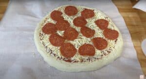 Homemade pepperoni pizza on parchment paper, ready to bake, with cheese and slices of pepperoni on a wooden surface.